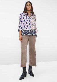 Donna con capelli lunghi e scuri che indossa una camicetta a maniche lunghe con fantasia geometrica, pantaloni larghi color taupe e stivali con tacco neri alla caviglia.