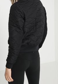 Svart vadderad bomberjacka med geometriskt mönster, ribbade ärmslut och en åtsittande nederkant. Tillverkad av lätt material för extra värme.