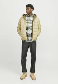 Chaqueta acolchada beige con capucha sobre una camisa a cuadros verde y blanca, combinada con jeans oscuros y botas de trabajo marrones.