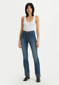 Jeans z wysokim stanem w niebieskim kolorze, lekko rozszerzane na dole, z wykończeniami szwów, pięcioma kieszeniami i zapięciem na zamek. Noszone z białym topem na ramiączkach.