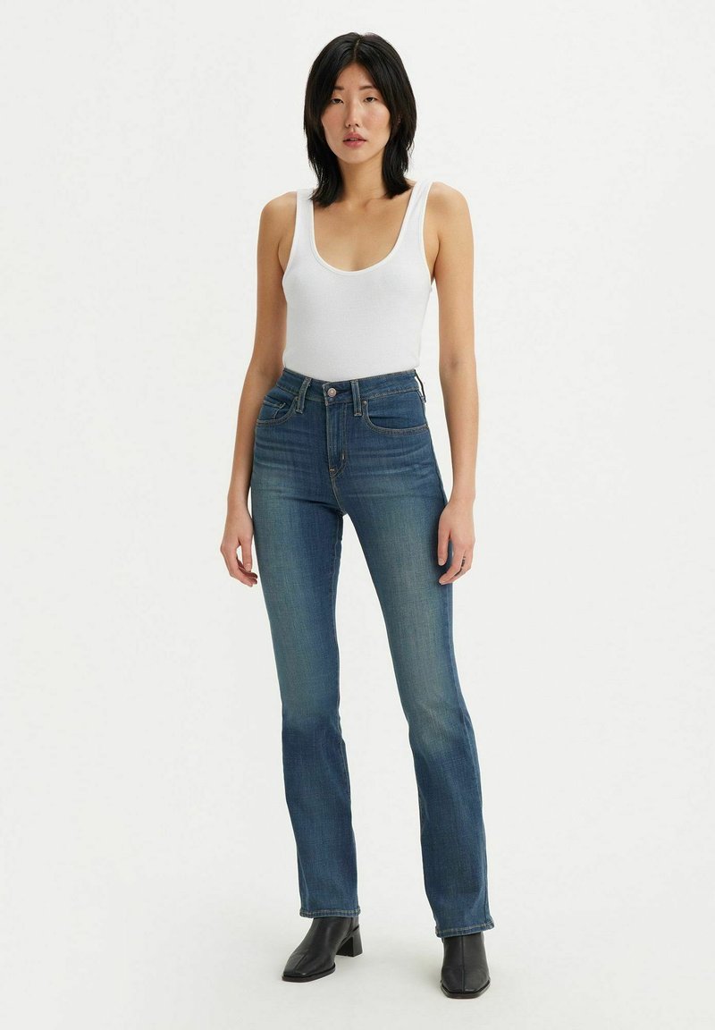 Jeans z wysokim stanem w niebieskim kolorze, lekko rozszerzane na dole, z wykończeniami szwów, pięcioma kieszeniami i zapięciem na zamek. Noszone z białym topem na ramiączkach.