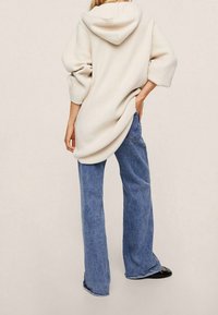 Beige gerippter übergroßer Kapuzenpullover mit einer Kapuze, kombiniert mit blauen weiten Jeans und schwarzen flachen Schuhen, von hinten betrachtet.