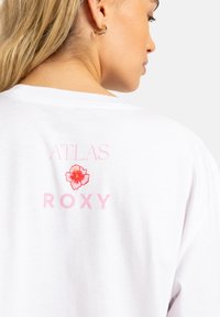 Hvit bomull T-skjorte med rosa "ATLAS" tekst over en blomsterdesign og "ROXY" tekst på baksiden. Myk tekstur, avslappet passform.