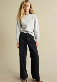 Grauer Sweatshirt mit überschnittenen Schultern, kombiniert mit hoch taillierten schwarzen Jeans, die ein dezentes Tigerstreifenmuster und weite Beine haben.