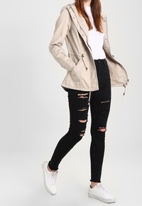 Beżowa kurtka z kapturem i sznurkami, w połączeniu z przetartymi czarnymi skinny jeansami i białymi sneakersami, na neutralnym tle.