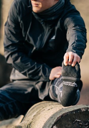 Schwarze Sportsocke mit strukturiertem Grip-Muster, versehen mit dem Logo "DANISH ENDURANCE". Getragen von einer Person in einer schwarzen Jacke, die auf einem Baumstamm sitzt.