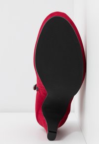 Botte cheville à talon haut en daim rouge avec semelle noire texturée et petit talon, positionnée verticalement contre un mur blanc d'angle.