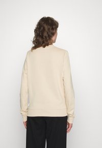 Persona con capelli ricci di lunghezza media, che indossa una felpa beige a maniche lunghe e pantaloni neri, in piedi di fronte a uno sfondo bianco.