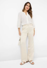 Blusa bianca a maniche lunghe con scollo a V, abbinata a pantaloni larghi color crema e sandali chiari, creando un look rilassato e stratificato.