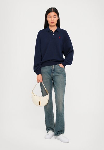 Femeie îmbrăcată în tricou polo bleumarin, blugi albaștri, adidași albi, ținând o mică poșetă albă, stând pe un fundal neutru.