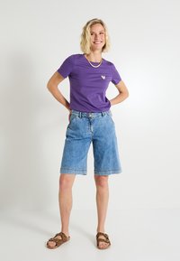 Camiseta morada de manga corta con un pequeño detalle de corazón dorado, combinada con pantalones cortos de mezclilla azul claro a la altura de la rodilla y sandalias marrones con estampado de leopardo.