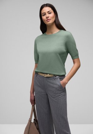 Femme portant un haut à manches courtes vert clair, un pantalon gris à fines rayures avec une ceinture beige, tenant un sac à main marron, posant devant un fond gris.
