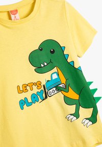 Gul kortærmet t-shirt med et grønt dinosaurusgrafik, der holder en blå lastbil, med teksten "LAD OS LEGE" i fede, farverige bogstaver.