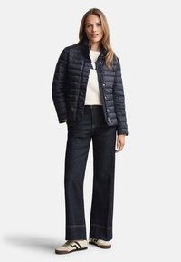 Marineblå dunjakke med quiltet tekstur, lynlås og trykknapper; kombineret med mørk denim flared bukser og cremefarvede sneakers med sorte accenter.