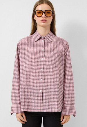 Giovane donna che indossa una camicia a quadri rosa oversize con bottoni e grandi occhiali da sole con lenti color ambra, in piedi davanti a uno sfondo semplice.