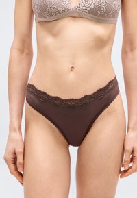 Γυναίκα που φοράει καφέ εσώρουχο με δαντέλα και αντίστοιχο μπεζ δαντελένιο σουτιέν, εμφανιζόμενη από τη μέση μέχρι τους μηρούς, στέκεται μπροστά σε απλό φόντο.