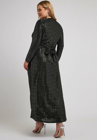 Zwarte langemouwen jurk met metalen zilveren strepen en een strik taille. Gemaakt van textuurstof, valt tot de enkels en bevat hoge sandalen.