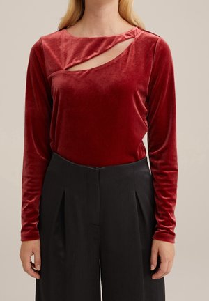 Kobieta ubrana w czerwony welurowy top z długim rękawem, z ukośnym wycięciem na klatce piersiowej, oraz czarne, plisowane spodnie z wysokim stanem, stojąca z rękami w luźnej pozycji.