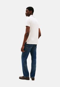 Camisa polo blanca, de mangas cortas, con cuello. Lleva puesta unos pantalones vaqueros azules y zapatos marrones. Diseño sencillo, sin patrones visibles.