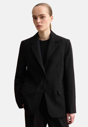 Vrouw met naar achteren gebonden haar, gekleed in een zwarte, op maat gemaakte blazer en een zwart topje, staand met één hand in de zak tegen een effen achtergrond.