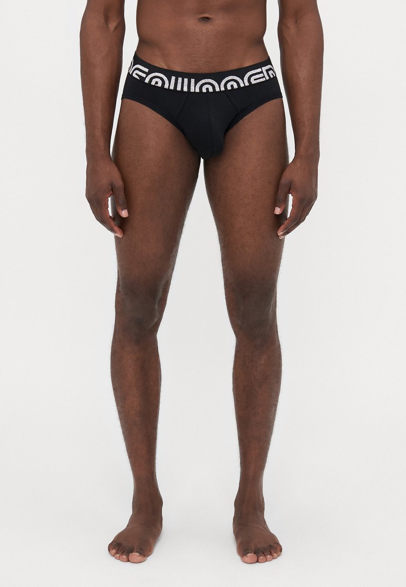 Sorte trusser med en tekstureret bæltedel, der har hvid skrift. Stoffet er glat med et tætsiddende design, der fremhæver et minimalistisk udseende.