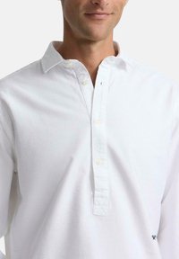 Camisa polo de manga larga de algodón blanco con tapeta de botones, cuello francés y pequeño emblema azul marino en el lado izquierdo.