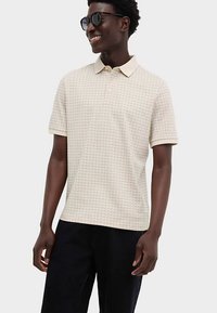 Polo beige à manches courtes avec un motif texturé, patte de boutonnage à trois boutons et col contrastant. Porté avec un pantalon foncé.