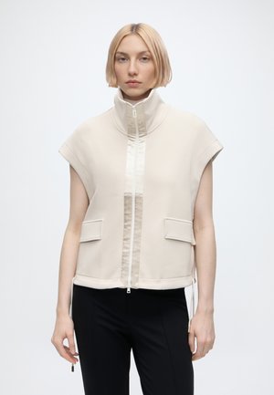 Femme blonde portant un gilet sans manches beige zippé avec col montant et poches à rabat, associé à un pantalon noir, debout devant un fond uni.
