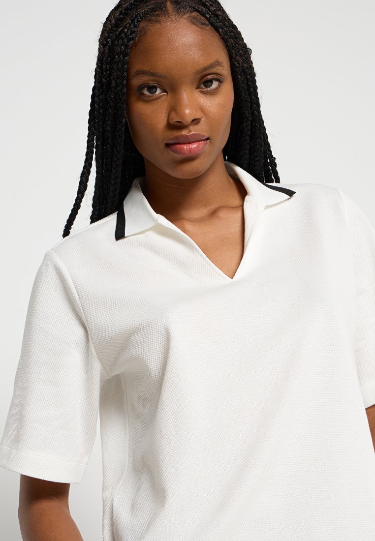 T-shirt polo blanc à manches courtes, présentant un tissu texturé et un accent noir sur le col. Design simple et épuré.