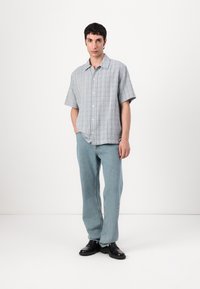 Kurzärmliges, hellblau gestreiftes Hemd mit Knopfverschluss, kombiniert mit lockeren hellblauen Jeans und schwarzen Schuhen. Einfarbiger Hintergrund.