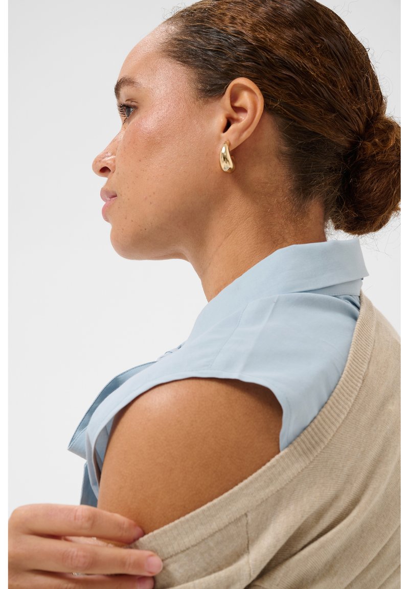 Femme à la peau brun clair et aux cheveux bouclés en chignon porte des boucles d'oreilles anneaux dorées, une chemise bleu clair à col et un pull en maille beige posé sur les épaules.