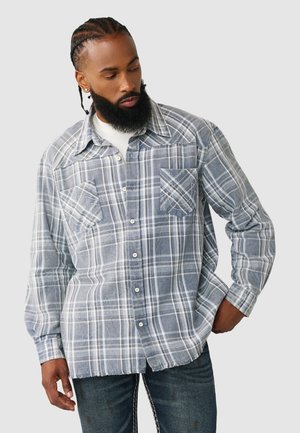 Mężczyzna z zaplecionymi włosami i brodą, ubrany w niebiesko-białą koszulę w kratę na guziki i ciemne dżinsy, patrzący w dół w lewo.