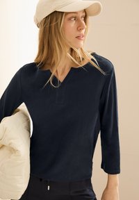 Marineblaue Langarmbluse mit V-Ausschnitt und strukturierter Stoff, getragen mit einer cremefarbenen, gesteppten Jacke, die einen lockeren Schnitt und einen lässigen Look bietet.