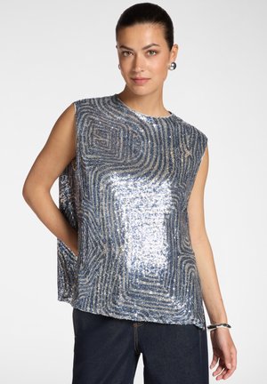 Vrouw met een mouwloze zilveren en blauwe pailletten top met geometrische patronen, donkere broek, zilveren oorbellen en een armband, staand met een hand in de zak.