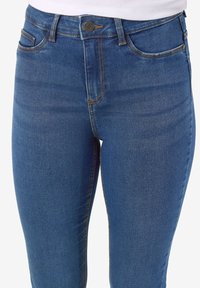Gros plan sur un jean en denim bleu avec poches avant, bouton et braguette à glissière porté avec un haut blanc.
