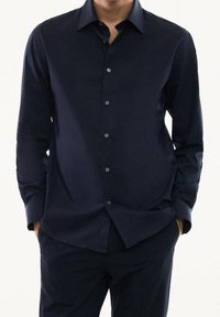 Chemise bleu marine à manches longues avec boutons, fabriquée en tissu lisse, avec un col pointu et des détails subtils de boutons sur le devant.