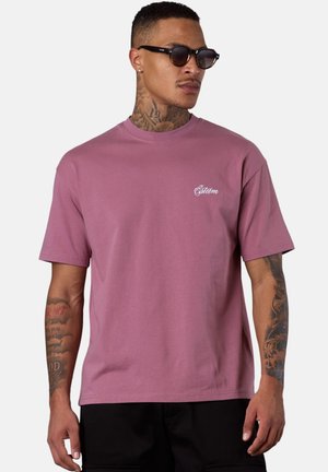 Mann mit Tattoos, der schwarze Sonnenbrille trägt, ein mauve-farbenes T-Shirt mit kurzen Ärmeln und kleinen weißen Schriftzügen sowie schwarze Hosen, leicht nach rechts gerichtet.