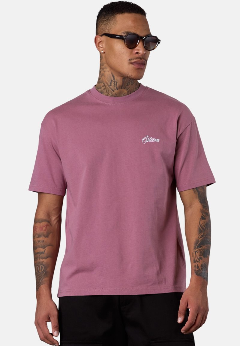 Mann mit Tattoos, der schwarze Sonnenbrille trägt, ein mauve-farbenes T-Shirt mit kurzen Ärmeln und kleinen weißen Schriftzügen sowie schwarze Hosen, leicht nach rechts gerichtet.