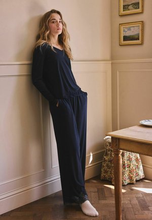 Woman leaning against a beige wall wearing a navy blue long-sleeve top and pants with white socks in a room with wood floor and floral stool.