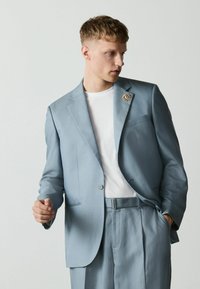 Jeune homme portant un costume bleu clair avec une chemise blanche et une épingle florale à la boutonnière, debout devant un fond clair uni, regardant vers la droite.