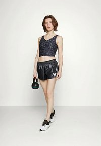 Top de desporto preto com um design estampado, combinado com calções pretos brilhantes e ténis brancos. A segurar um kettlebell preto.