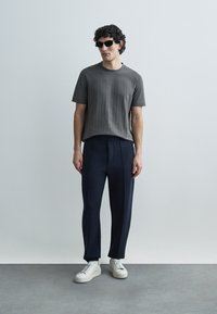 Grey textured short-sleeve shirt, dark navy pants with a tailored fit, and white sneakers. Model wears black sunglasses.