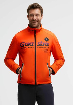 Man wearing bright orange zip-up jacket with hands in pockets, smiling, standing against plain light background.