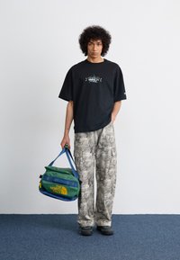 Camiseta grande negra con diseño gráfico, pantalones cargo de camuflaje claro y una bolsa de deporte en verde y azul con logo amarillo. Zapatos negros.