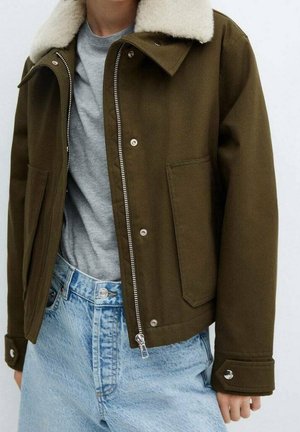 Person trägt eine olivgrüne Jacke mit weißem Schafsfellkragen, graues Hemd und hellblaue hoch taillierte Jeans mit Reißverschluss.