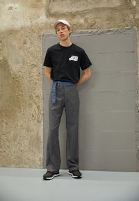 Camiseta preta com logótipo, calças cinzentas de alta cintura, cinto de ganga azul. Modelo veste sapatilhas pretas e está encostado a uma parede texturizada.