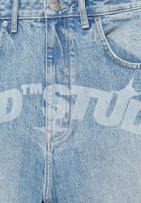 Jean en denim bleu clair avec poches avant, coutures visibles, un bouton en métal et texte blanc imprimé délavé sur le tissu.