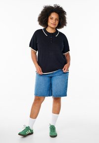 Svart pikétröja med kontrasterande krämfärgad krage och manschetter, tillsammans med ljusblå denimshorts. Modellen har på sig gröna sneakers och vita strumpor.
