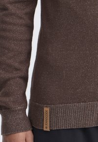 Brauner Pullover mit strukturiertem Strickmuster, ausgestattet mit einem gerippten Saum. Enthält ein hellbraunes Etikett mit "JEANSCODE". Subtile Funken sind im Stoff sichtbar.