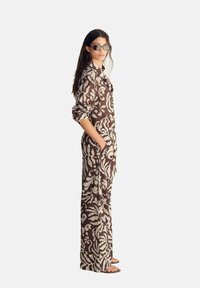 Brun mønstrede jumpsuit med blomsterdesign, lange ærmer og lommer, parret med sorte solbriller og sandaler. Glat stoftekstur.